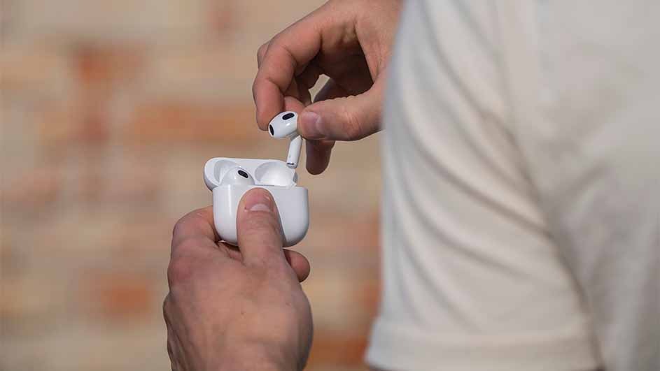 A person holds a white earbud case, removing one earbud; another remains in the open case. A white shirt and brick wall backdrop.