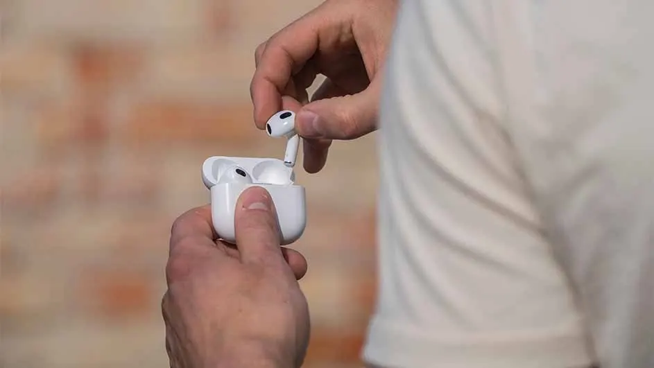 A person holds a white earbud case, removing one earbud; another remains in the open case. A white shirt and brick wall backdrop.