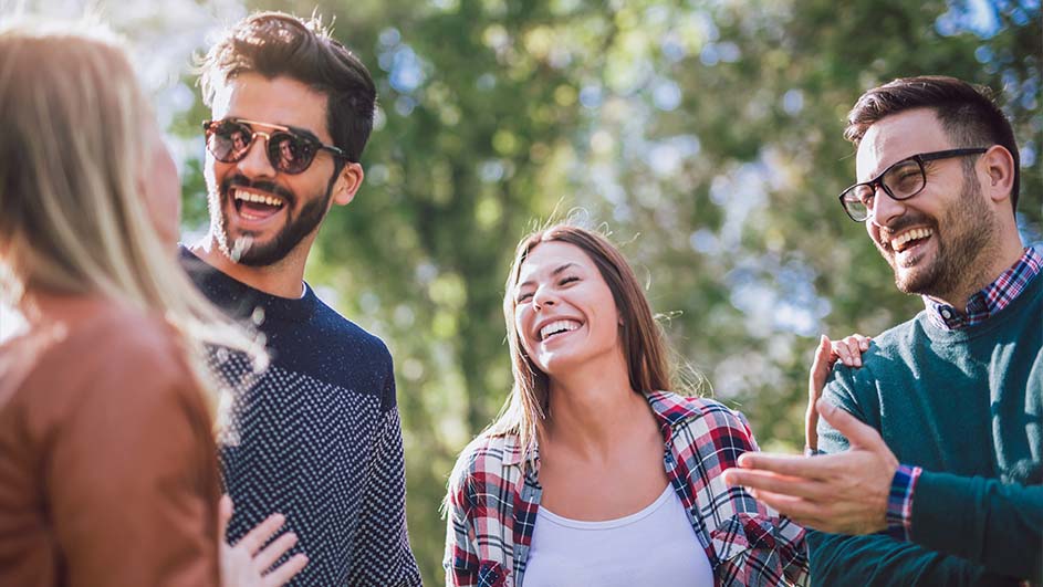 Vier lachende mensen staan buiten. Twee mannen dragen een bril, een vrouw een geruite blouse. Groene bomen op de achtergrond.