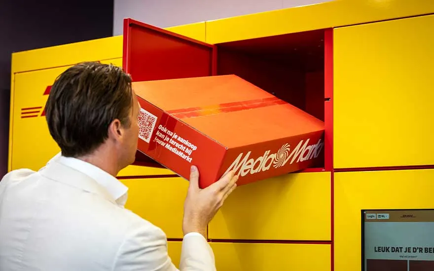 Een man in wit haalt een rode MediaMarkt-doos uit een felgele DHL-pakjeskluis, klaar om zijn bestelling op te halen.