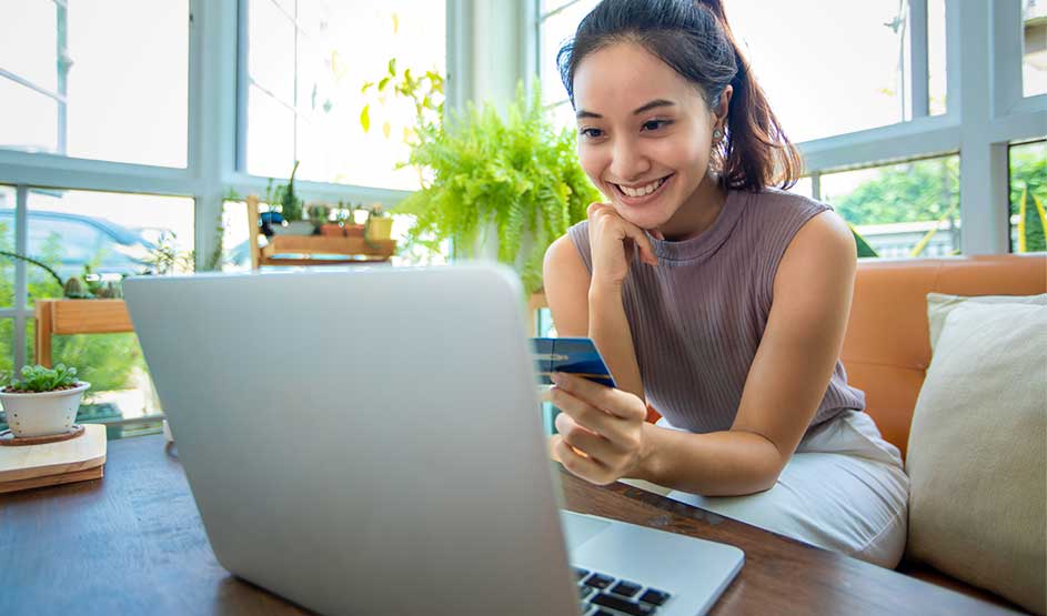 Glimlachende vrouw met creditcard kijkt naar laptop in lichte kamer met planten en grote ramen.