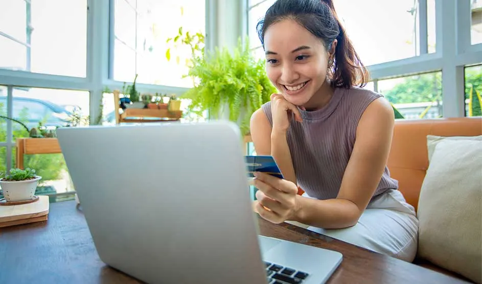 Glimlachende vrouw met creditcard kijkt naar laptop in lichte kamer met planten en grote ramen.