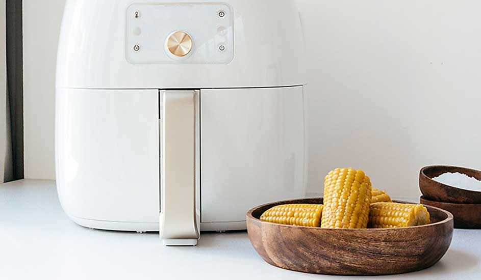 Een witte airfryer staat op het aanrecht met daarnaast een houten kom met gekookte maiskolven. Het merk van de airfryer is niet zichtbaar.