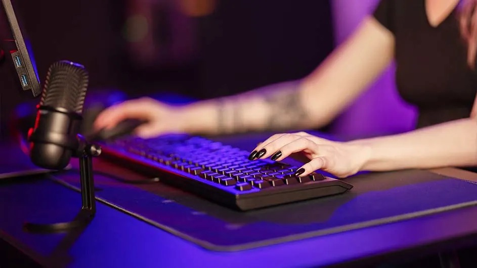 A person with a tattoo is using a black computer keyboard and mouse. A microphone stands to the left on a dark blue desk.