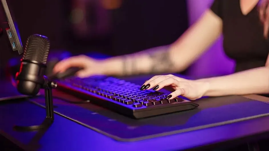 A person with a tattoo is using a black computer keyboard and mouse. A microphone stands to the left on a dark blue desk.