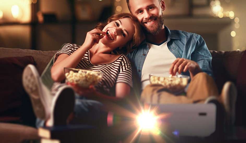 A couple sits close together on a cosy sofa, each holding a bowl of popcorn. A projector beams light towards the screen. No brand name is visible.