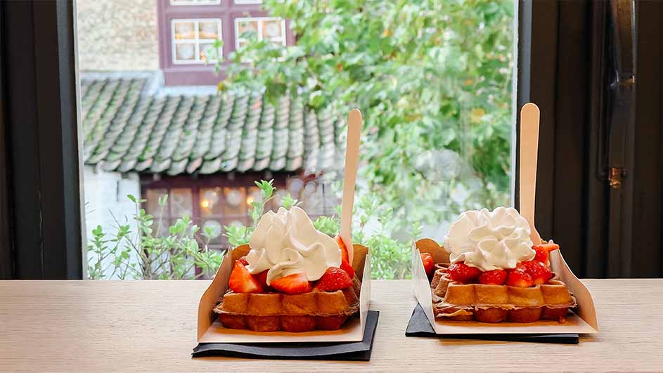 Twee wafels, elk met aardbeien en slagroom, staan op een tafel voor een raam met uitzicht op een gebouw.