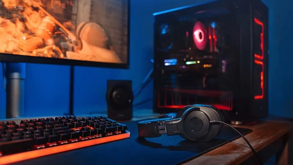 Gaming setup with lit keyboard, headphones on the desk, and a glowing PC tower beside a monitor showing an action scene.