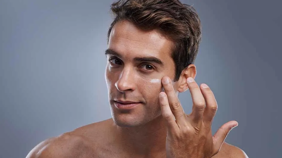Een man smeert witte crème onder zijn oog met zijn rechterhand tegen een blauwgrijze achtergrond.