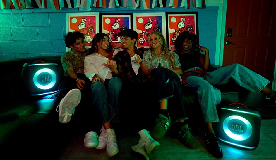 Vrienden zitten dicht bij elkaar op een bank, lachend in groen licht, terwijl twee ronde JBL-speakers naast hen helder oplichten.