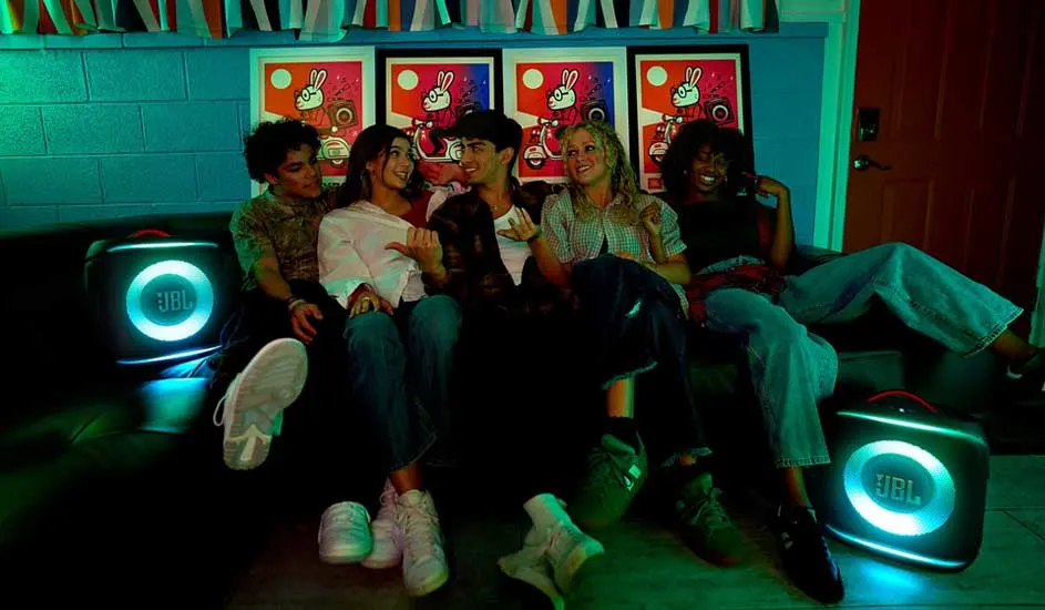 Vrienden zitten dicht bij elkaar op een bank, lachend in groen licht, terwijl twee ronde JBL-speakers naast hen helder oplichten.