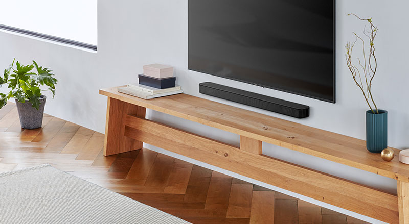 Een modern ingerichte woonkamer met lichtvisgraatvloer, aan de muur een grote flatscreen-tv met daaronder een zwarte soundbar op een robuuste houten bank, links een stapel boeken en een groene kamerplant.