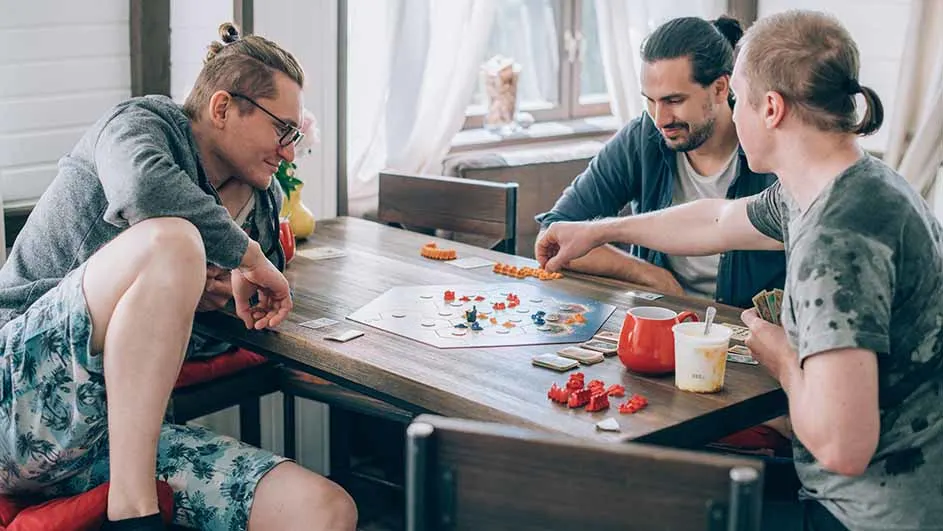 Drie mannen spelen een bordspel aan een houten tafel met kaarten en drankjes in een kamer met een raam.