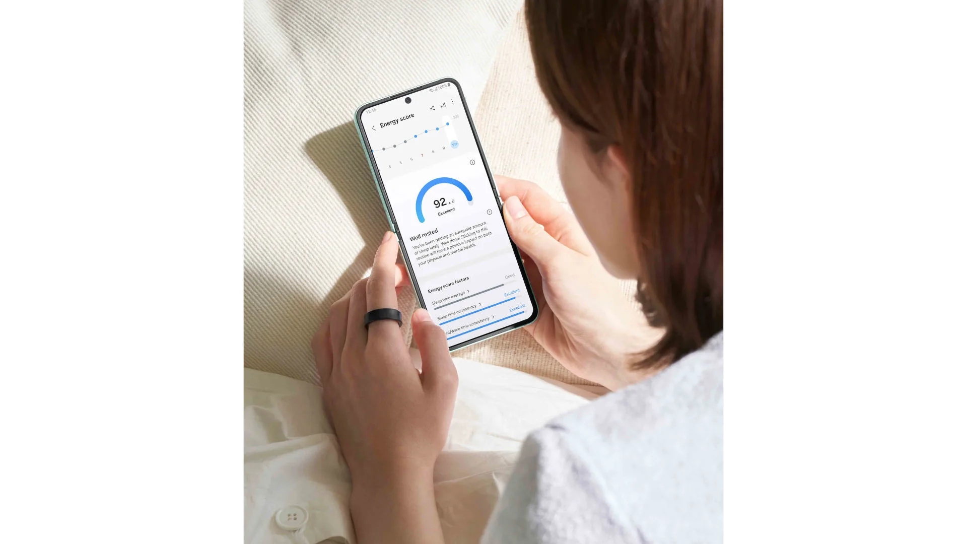 Vrouw bekijkt energie-score (92.0) op een smartphone, met een zwarte ring om haar vinger, zittend op een crèmekleurige ondergrond.