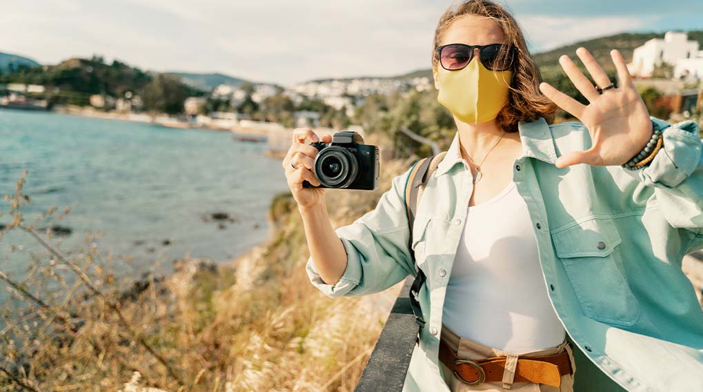 Een vrouw met een geel masker houdt een camera vast, met zee en gebouwen op de achtergrond.
