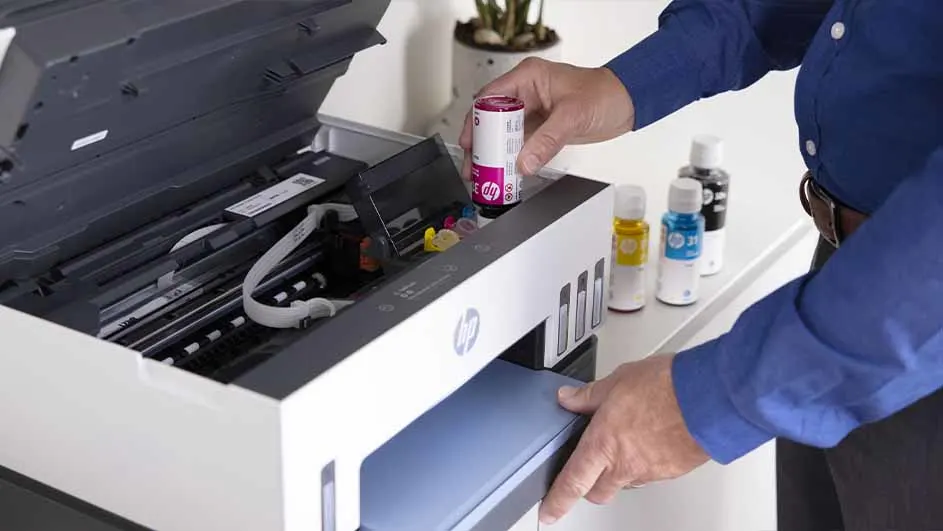 Een persoon vult een witte HP printer met een roze inktpatroon; flessen gele, blauwe en zwarte inkt staan klaar.