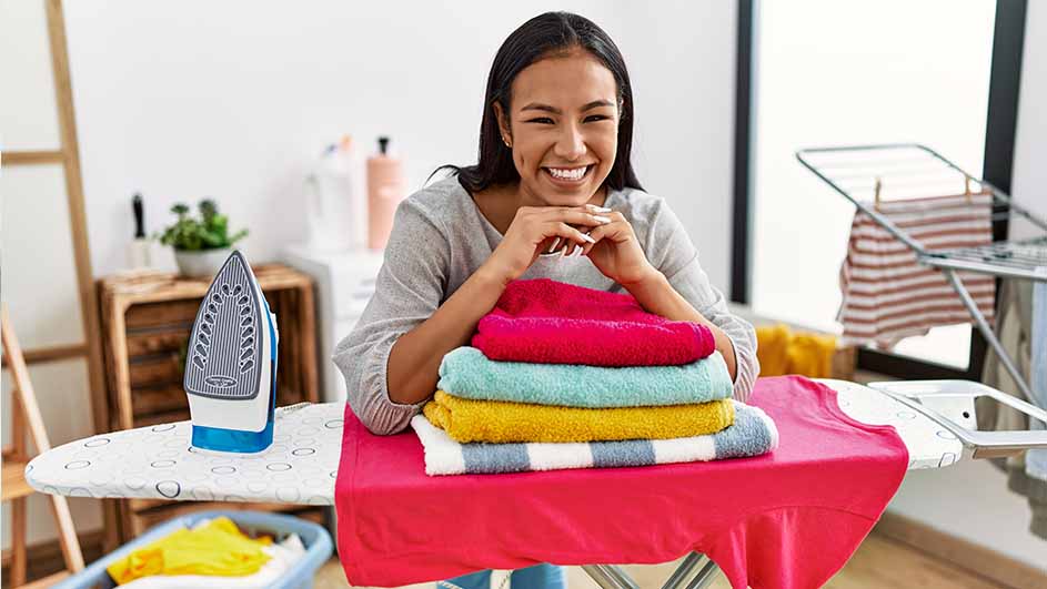 Vrouw glimlacht, leunt op gestapelde kleurrijke handdoeken op strijkplank. Strijkijzer, wasrek en wasmand zichtbaar.