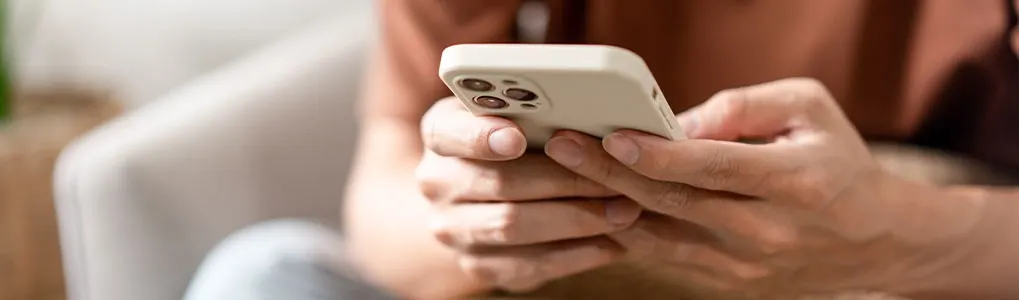 A person holds a white smartphone with multiple lenses, thumbs on the screen. Background: a light-colored upholstered chair.