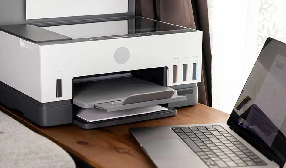 Een witte printer met gekleurde inktreservoirs staat naast een opengeklapte laptop op een houten tafel. Merk is niet zichtbaar.