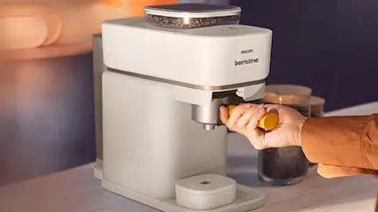 Een Philips Baristina-koffiezetapparaat met een hand die een gele hendel bedient, op een aanrecht naast een glazen pot.