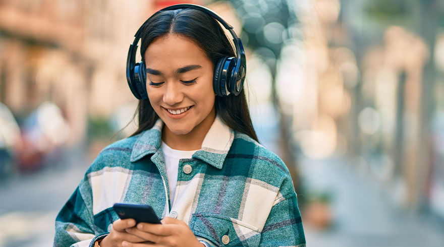Jonge vrouw met koptelefoon en geruit overhemd kijkt lachend naar een telefoon. Achtergrond is vaag en toont een stedelijke omgeving.