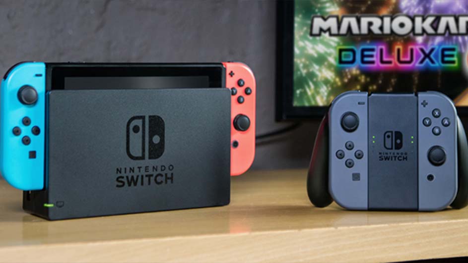 Een Nintendo Switch-console met blauwe en rode Joy-Cons in het dock, plus een controller op een houten tafel. "Mario Kart Deluxe" op scherm.