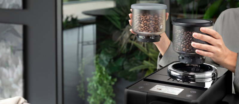 Twee handen plaatsen koffiebonenreservoirs op een zwart koffiezetapparaat. Achtergrond toont een raam en groene planten.