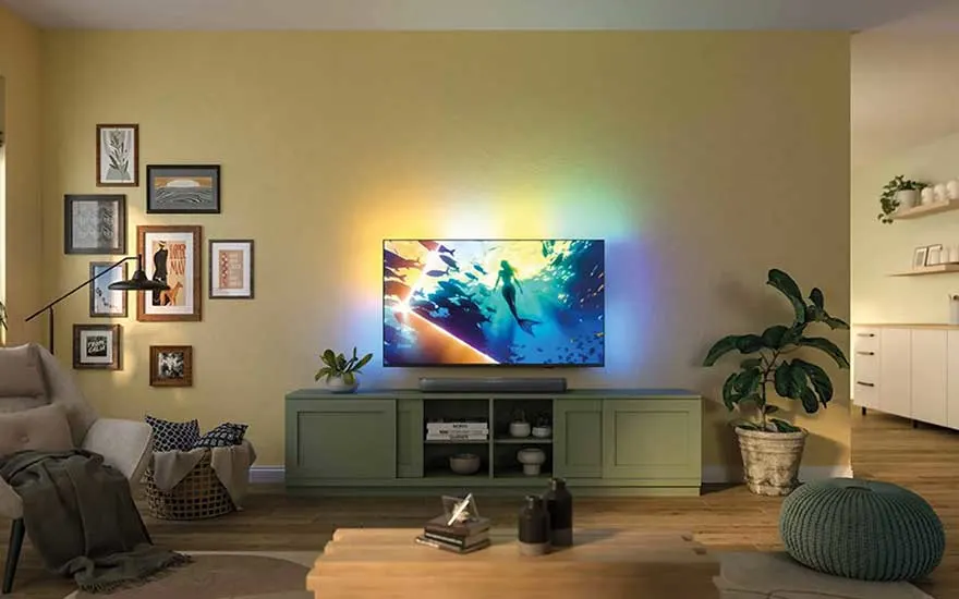 In a cosy living room, a TV sits on a green cabinet, colourful lights glow behind it, and plants bring warmth to the space.