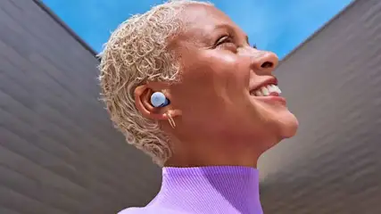 Een vrouw met kort blond haar draagt Pixel Buds en glimlacht breed, terwijl ze naar de lucht kijkt onder felle zon.