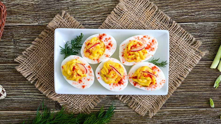 Zes gevulde eieren op een wit bord, gegarneerd met groene kruiden en rode paprika, op een rustieke houten tafel met jute.