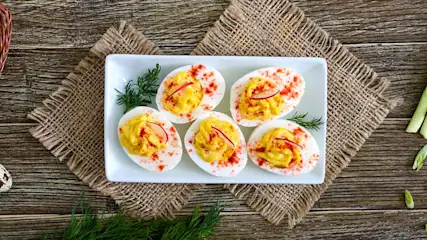 Zes gevulde eieren op een wit bord, gegarneerd met groene kruiden en rode paprika, op een rustieke houten tafel met jute.