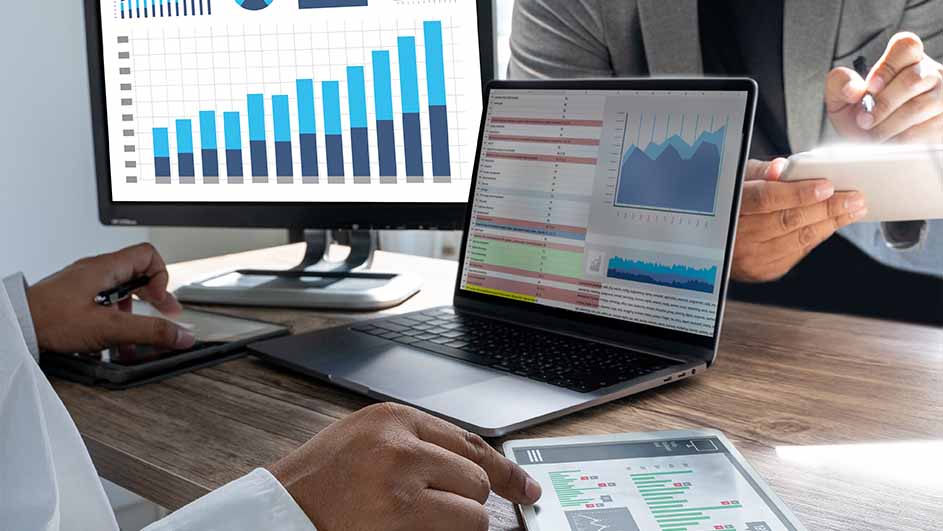 Twee personen werken aan een houten bureau met een laptop, tablet en monitor die grafieken weergeven.