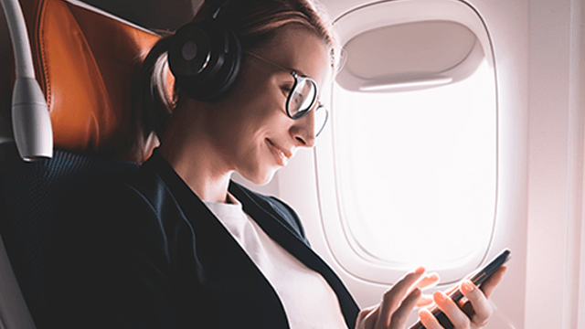 Een vrouw met een koptelefoon en een bril zit in een vliegtuig en gebruikt een telefoon. Het vliegtuigraam is helder.