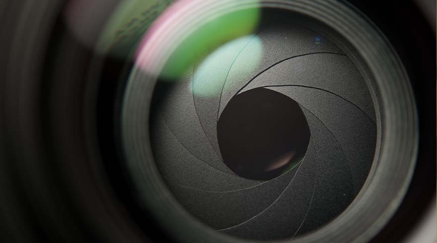 Close-up van een grijze cameralens met een opening in het midden, omgeven door reflecties van groen en roze licht.