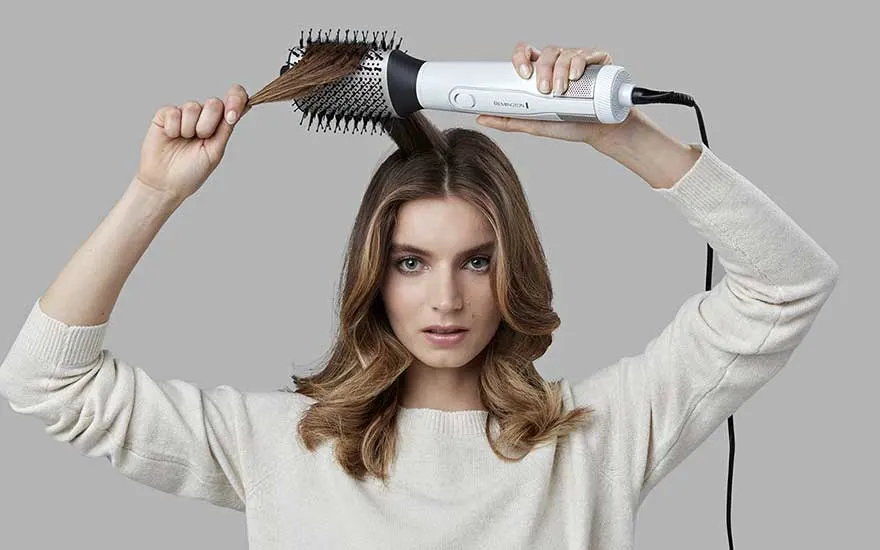 Vrouw met halflang blond haar gebruikt een witte warme luchtborstel bovenop haar hoofd, terwijl zij met de hand een haarlok strak rond de borstel trekt.