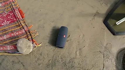 Een blauwe JBL-speaker staat op zand, naast een rood gestreept kleed met een steen erop. Een zwart frame is rechts zichtbaar.