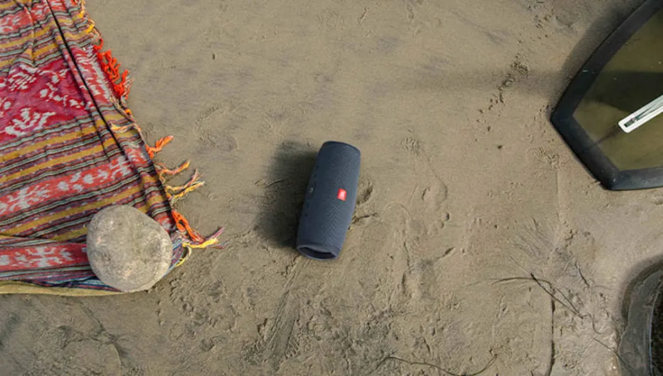 Een blauwe JBL-speaker staat op zand, naast een rood gestreept kleed met een steen erop. Een zwart frame is rechts zichtbaar.