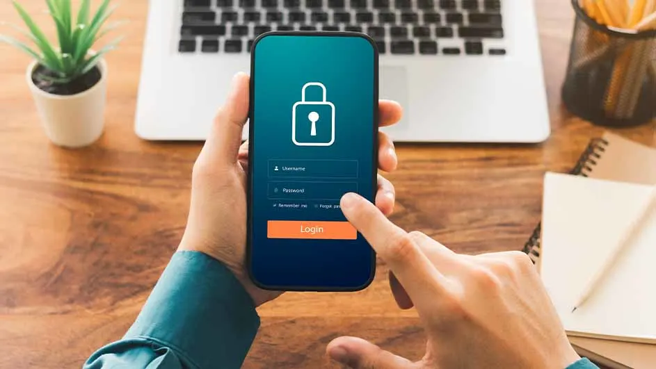 Handen houden een telefoon met een blauw scherm met een slotpictogram en een login-knop, op een houten bureau met een laptop.
