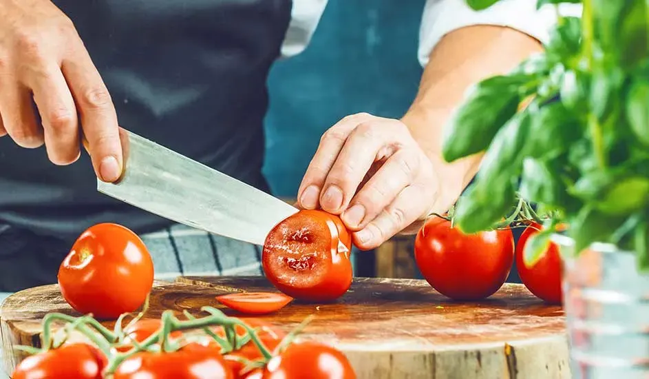Een persoon snijdt rode tomaten op een houten plank met een zilveren mes, basilicumplant op de achtergrond.
