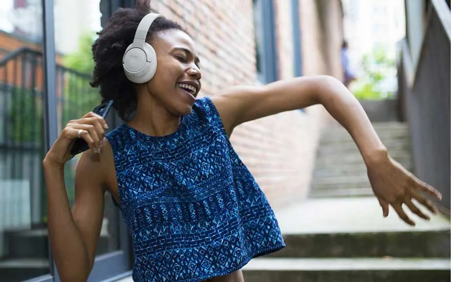 Woman with white headphones dances joyfully in an alley outdoors, phone in hand, completely absorbed in the music.