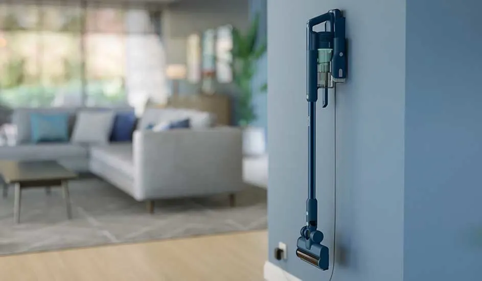 A slim blue stick vacuum hangs on the wall of a modern living room, always charged and ready to pick up crumbs and dust.