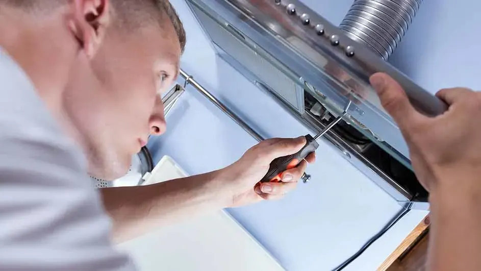 Installer carefully fixing a cooker hood, using a screwdriver while a flexible metal vent duct runs above the unit.