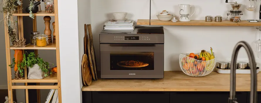 A kitchen features an oven with pizza inside, a fruit basket, a sink, and a wooden shelf with various items.