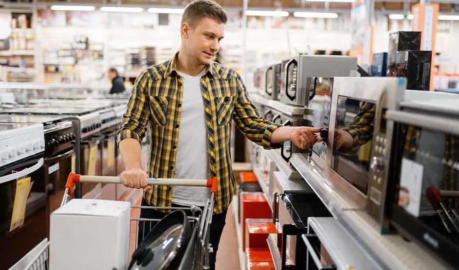 Een man in een geruit overhemd bekijkt een magnetron in een elektronicawinkel en duwt een winkelwagen vol apparaten. Geen merk zichtbaar.