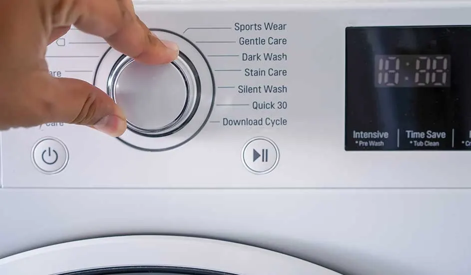 A hand is turning the programme dial of a washing machine. Various washing modes are labelled next to the dial. No brand name is visible on the appliance.
