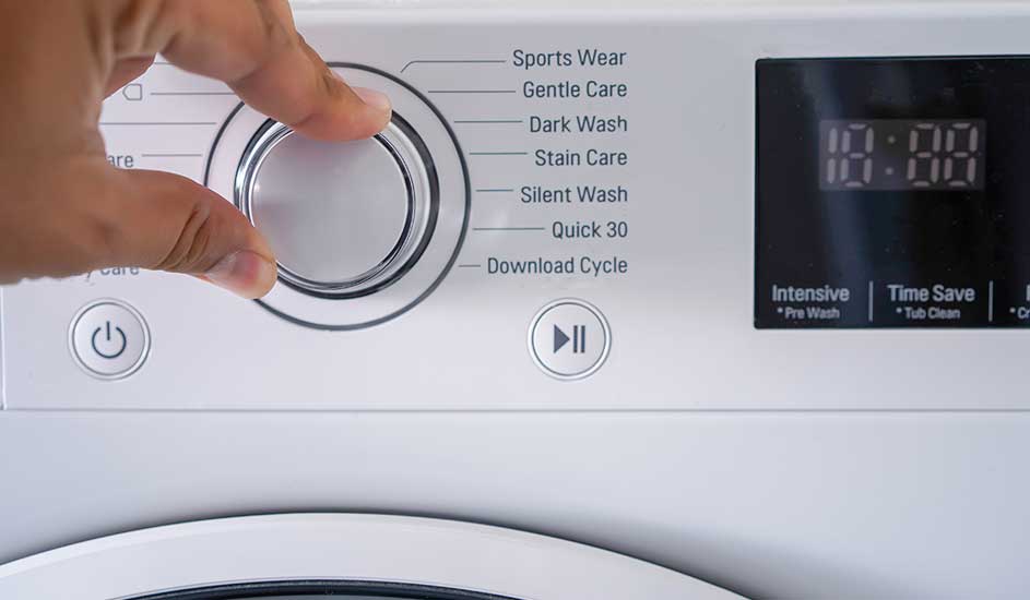 Een hand draait aan de programma-knop van een wasmachine. Op het display staan diverse wasprogramma’s en een digitaal scherm.