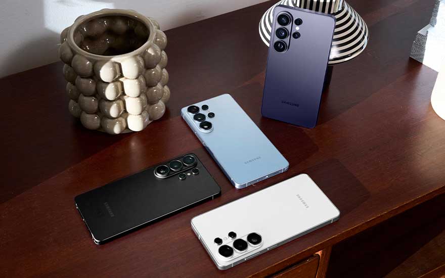 Vier Samsung smartphones in verschillende kleuren liggen op een houten dressoir naast een opvallende keramische vaas en een zwart wit gestreept object.