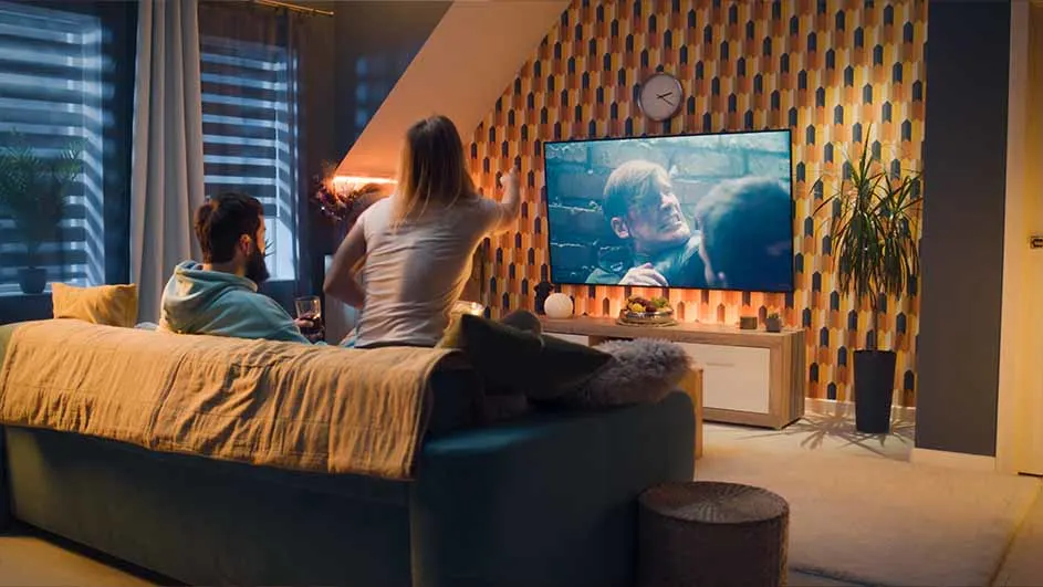 Woonkamer met een man en vrouw op een blauwe bank voor een tv met een film aan. Een plant staat naast de tv.