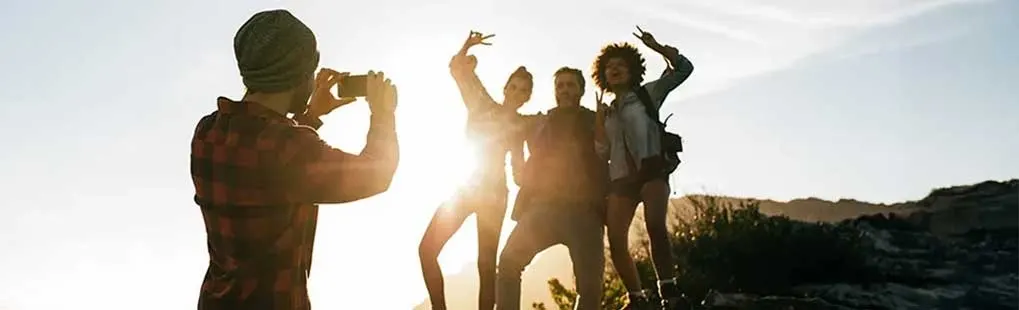 A person in a hat and red shirt takes a photo of four friends on a rocky peak against a bright sky.