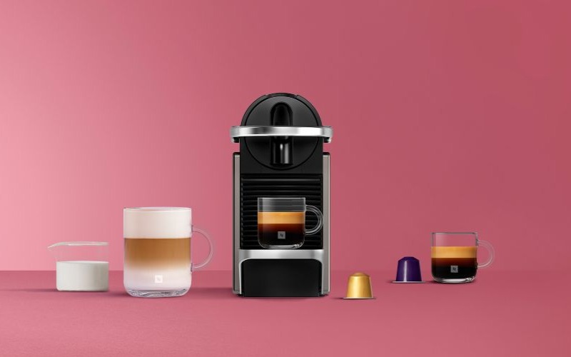 Zwarte Nespresso koffiemachine met twee glazen kopjes koffie en kleurrijke capsules op een roze achtergrond.